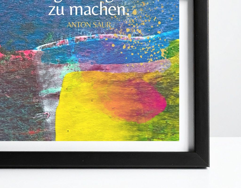 Schwarzer Bilderrahmen mit einem farbenfrohen, abstrakten Kunstwerk, das hauptsächlich blaue, gelbe, pinke und grüne Farbtöne zeigt, mit einem Teil eines Zitats von Anton Saur.